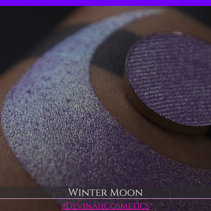 A close-up of a person's arm with a large purple eyeshadow swatch and a matching circular hand made eyeshadow pan, labeled 'Winter Moon' and '#DevinahCosmetics" 