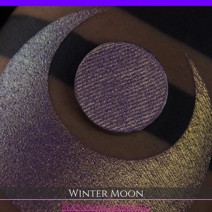 A close-up of a person's arm with a large purple eyeshadow swatch and a matching circular hand made eyeshadow pan, labeled 'Winter Moon' and '#DevinahCosmetics" 