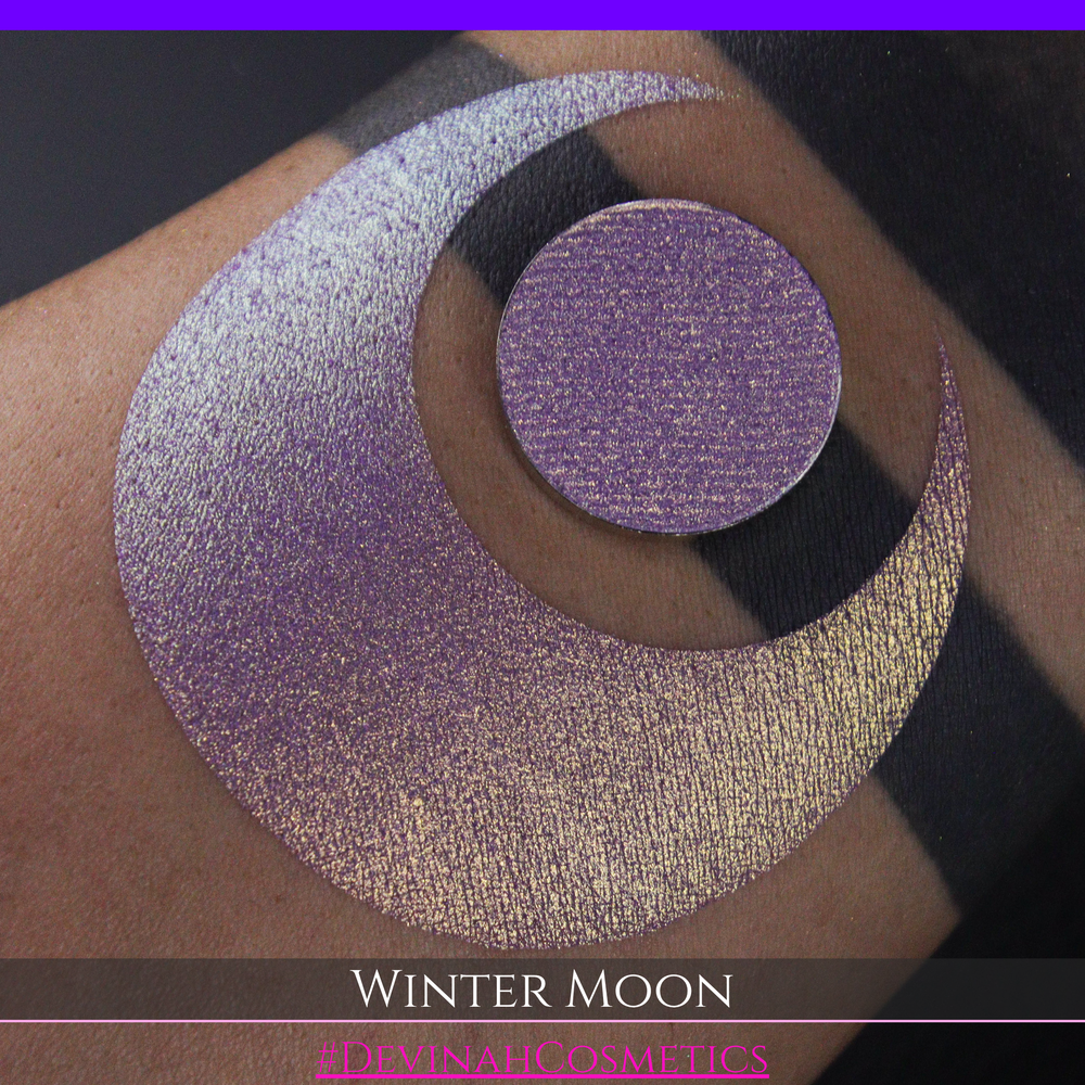 A close-up of a person's arm with a large purple eyeshadow swatch and a matching circular hand made eyeshadow pan, labeled 'Winter Moon' and '#DevinahCosmetics" 