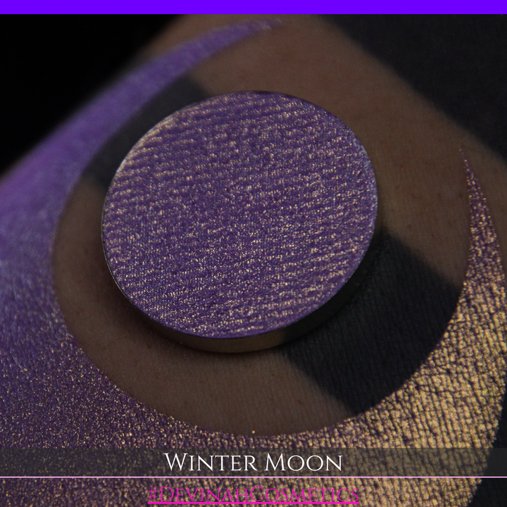 A close-up of a person's arm with a large purple eyeshadow swatch and a matching circular hand made eyeshadow pan, labeled 'Winter Moon' and '#DevinahCosmetics" 