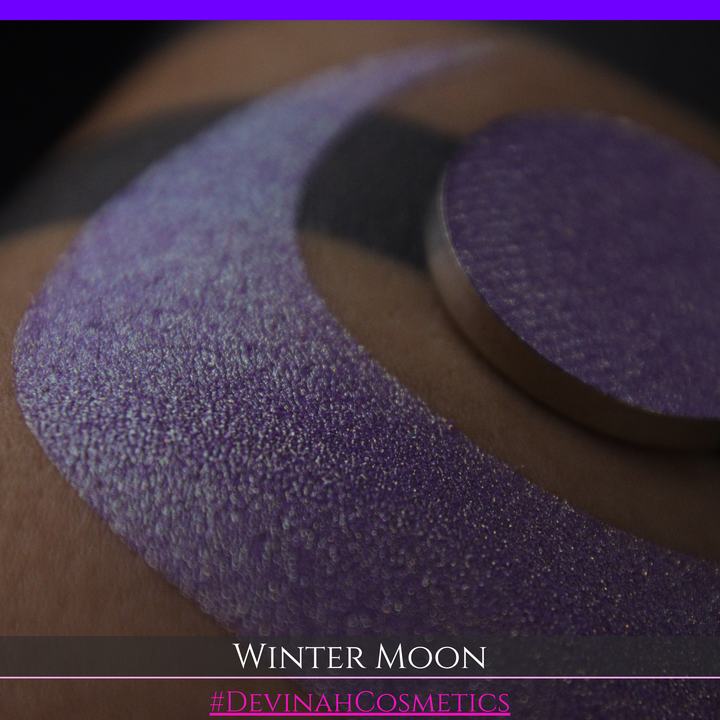 A close-up of a person's arm with a large purple eyeshadow swatch and a matching circular hand made eyeshadow pan, labeled 'Winter Moon' and '#DevinahCosmetics" 