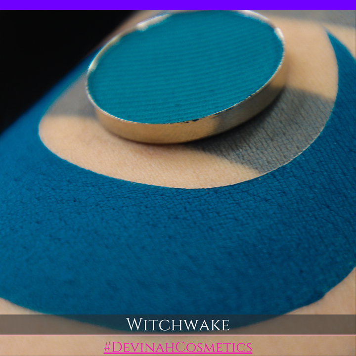 A close-up of a circular teal eyeshadow pan resting on top of a swatch of blue and beige eyeshadow, with the text 'WITCHWAKE' and '#DEVINAHCOSMETICS' at the bottom.