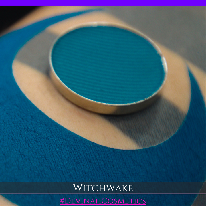 A round teal eyeshadow pan with a gold-toned metal rim, resting on a textured blue swatch. Text at the bottom reads 'WITCHWAKE' and '#DEVINAHCOSMETICS'.