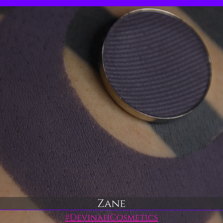 A makeup swatch showing a dark plum-purple eyeshadow color with a circular makeup pan and the name 'ZANE' in white text on a black background.