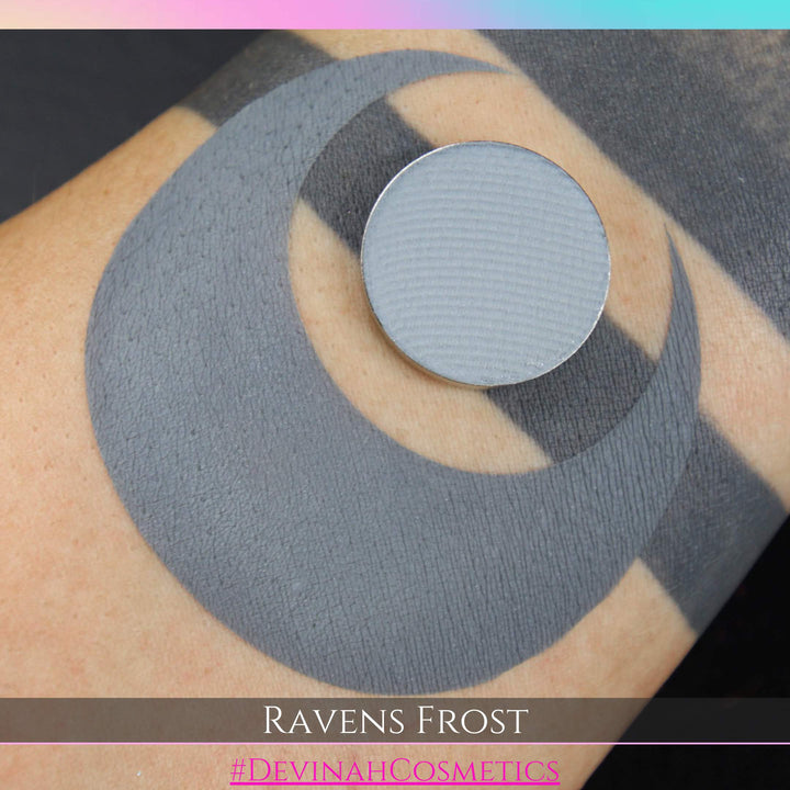 Close-up of a circular eyeshadow pan with a swatch on tan skin with 'Ravens Frost' and 'Devinah Cosmetics' branding.